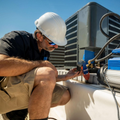 Technician in a hard hat kneels beside rooftop HVAC equipment, inspecting wiring under a clear blue sky