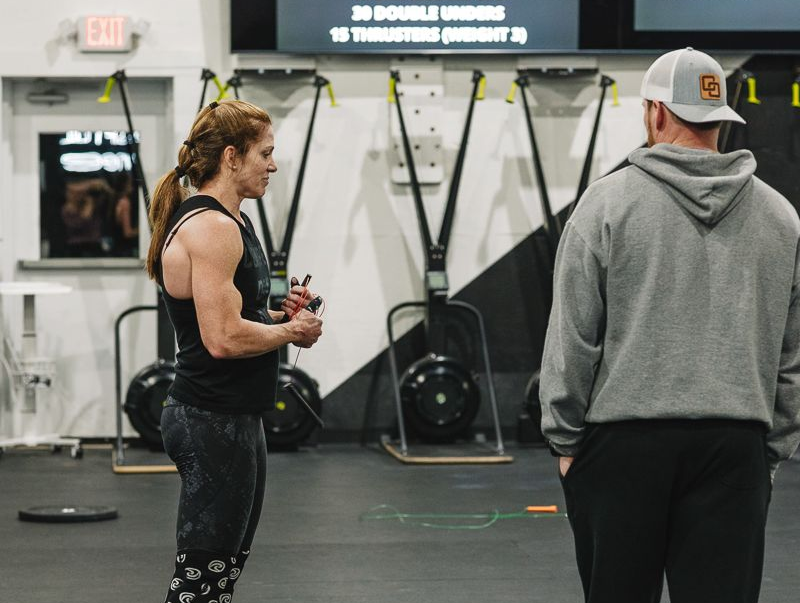 Woman in workout gear talking to a man in a gym, with exercise equipment in the background.