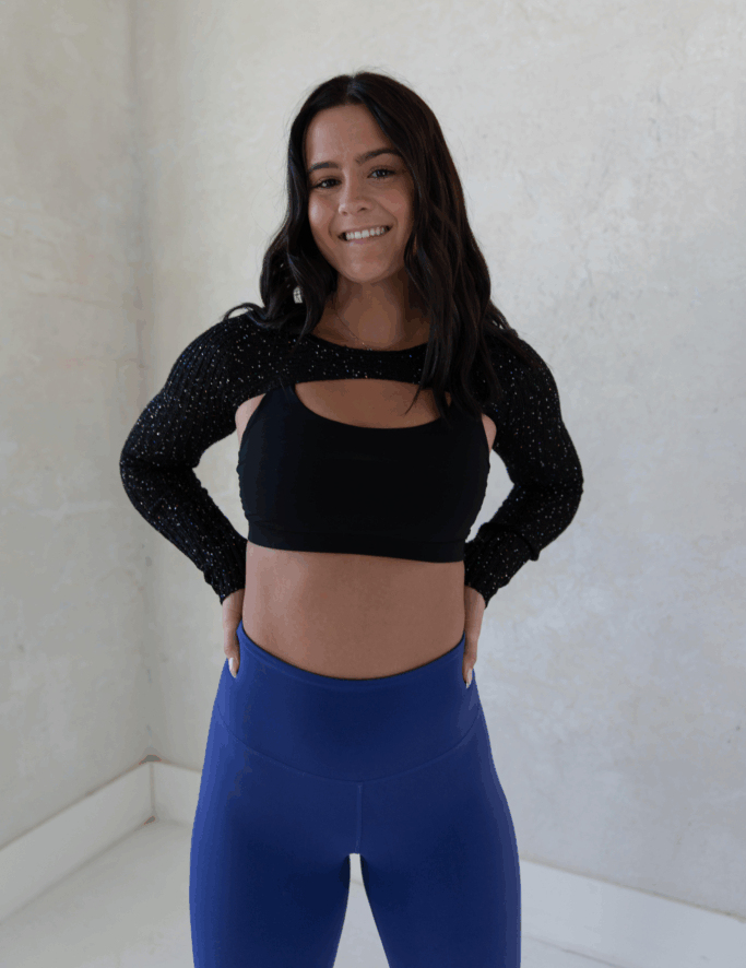 Woman smiling, wearing black cutout top with sparkly sleeves, blue leggings, hands on hips. White wall backdrop.
