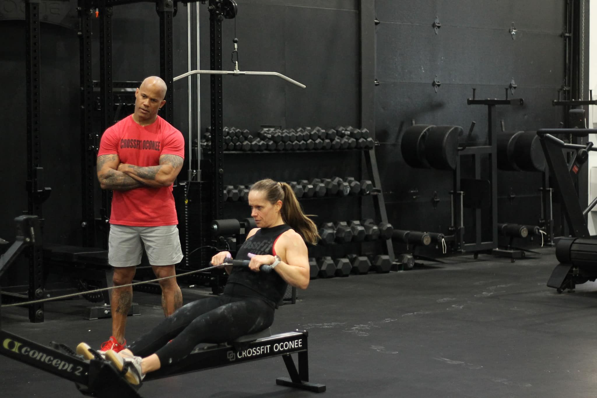 Woman rowing on a Concept2 machine, instructor watches. Gym setting.