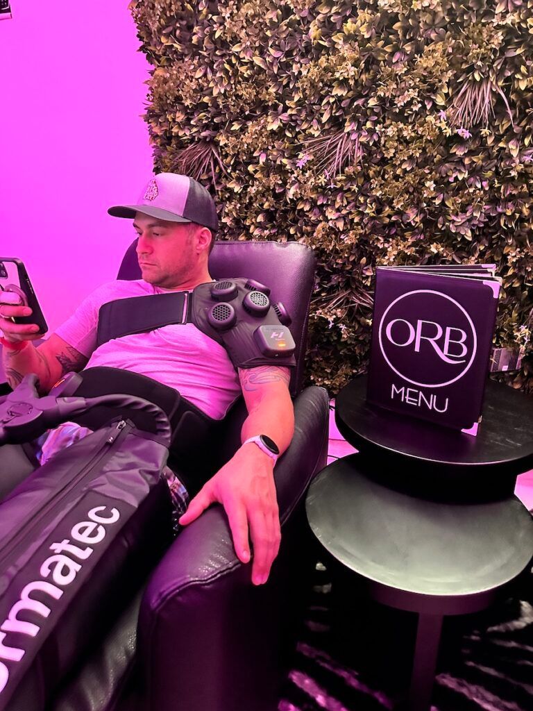 Man in recovery chair using phone, connected to compression sleeves. ORB menu on table, green wall.