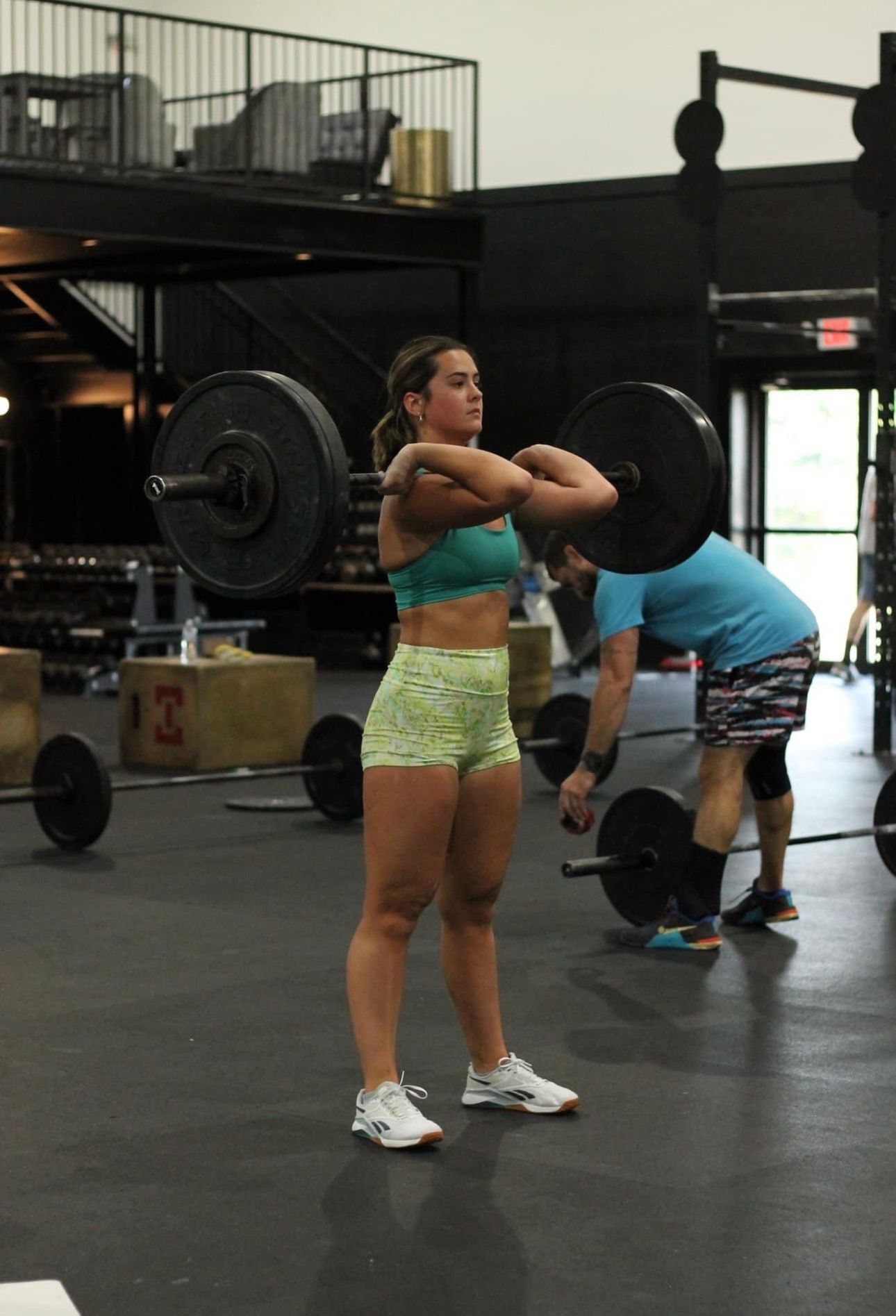 Woman in gym holding barbell at shoulder level, wearing athletic wear. Another person visible in background.