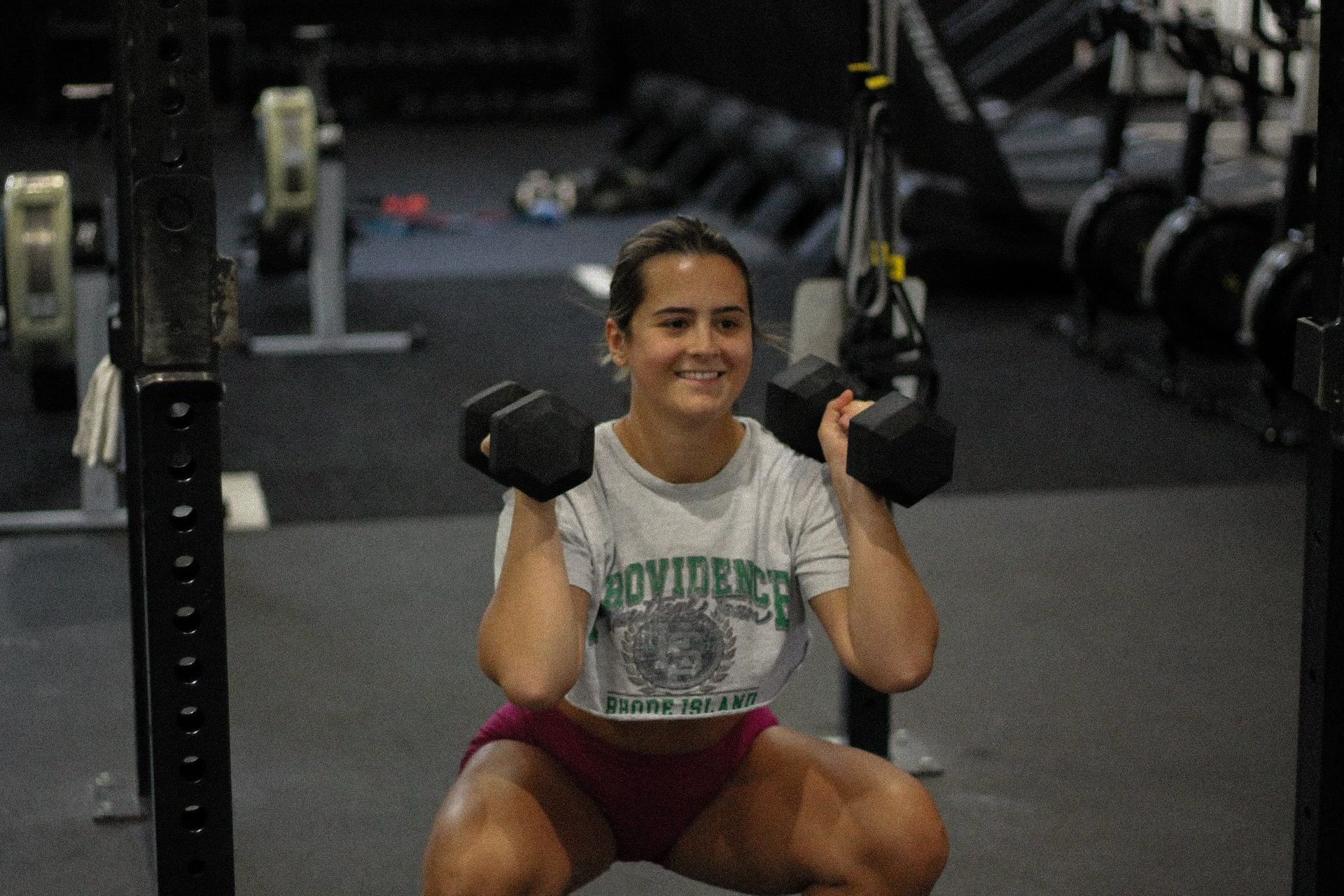 Woman in a white shirt squats while holding dumbbells in a gym, smiling.