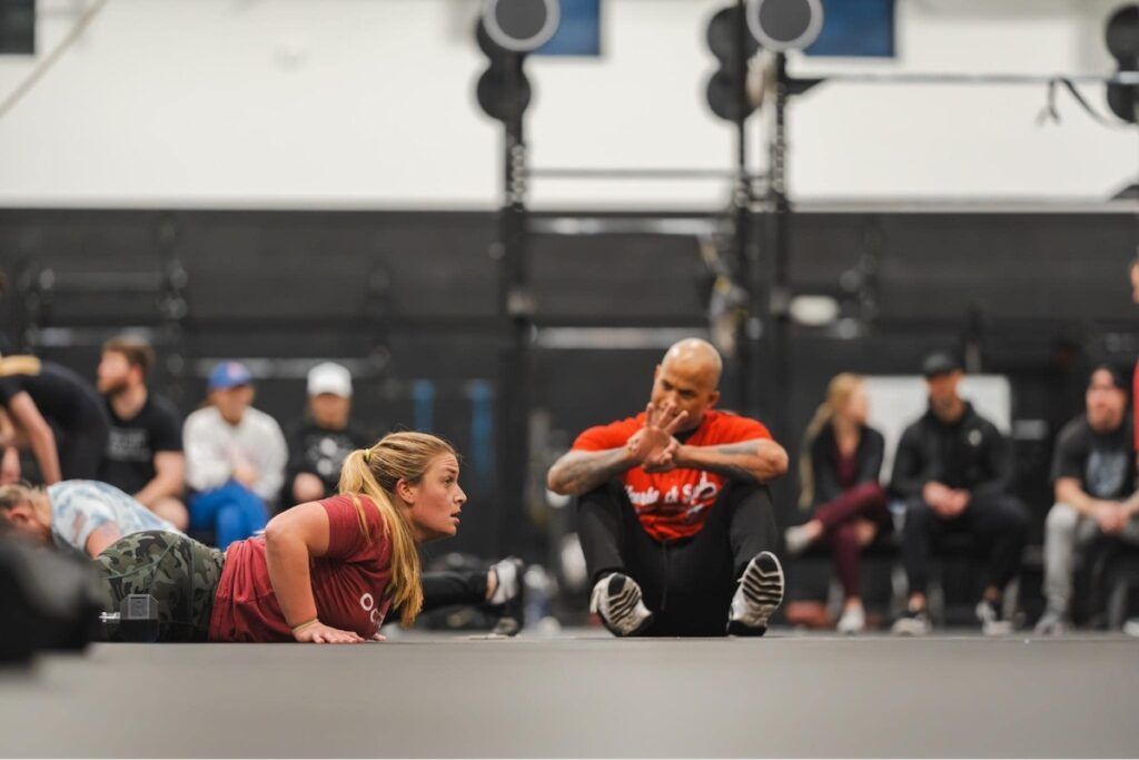 Woman doing push-ups on floor, watched by coach in gym. People seated in background.