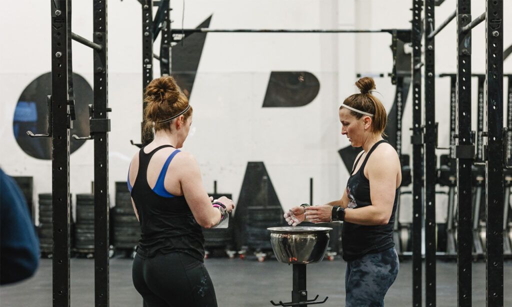 Two women in a gym, preparing for a workout, one with chalk. Workout rig and logo in the background.