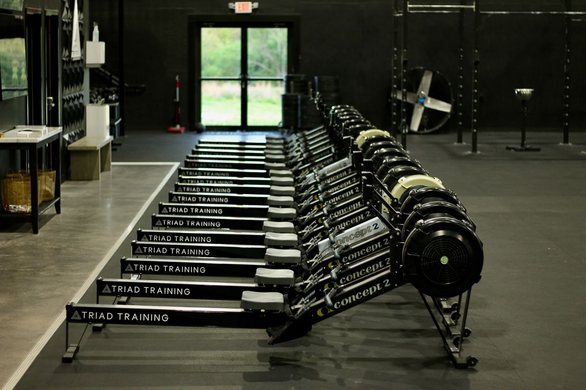 Row of Concept2 rowing machines in a gym, facing a window and an exit sign.