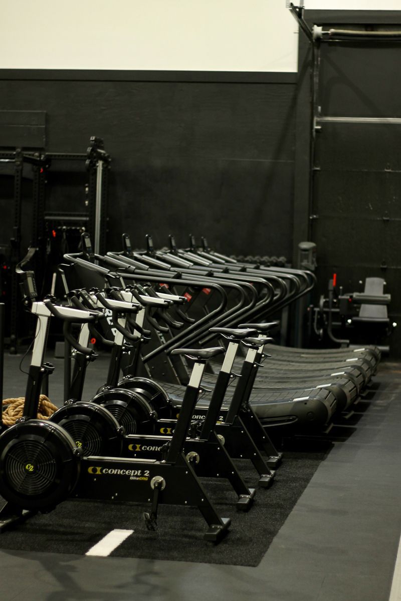 Row of stationary exercise bikes, treadmills, and rowing machines in a dark gym.