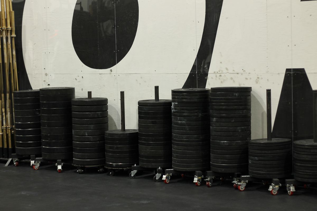 Black weight plates stacked on rolling carts in a gym, against a white and black wall.