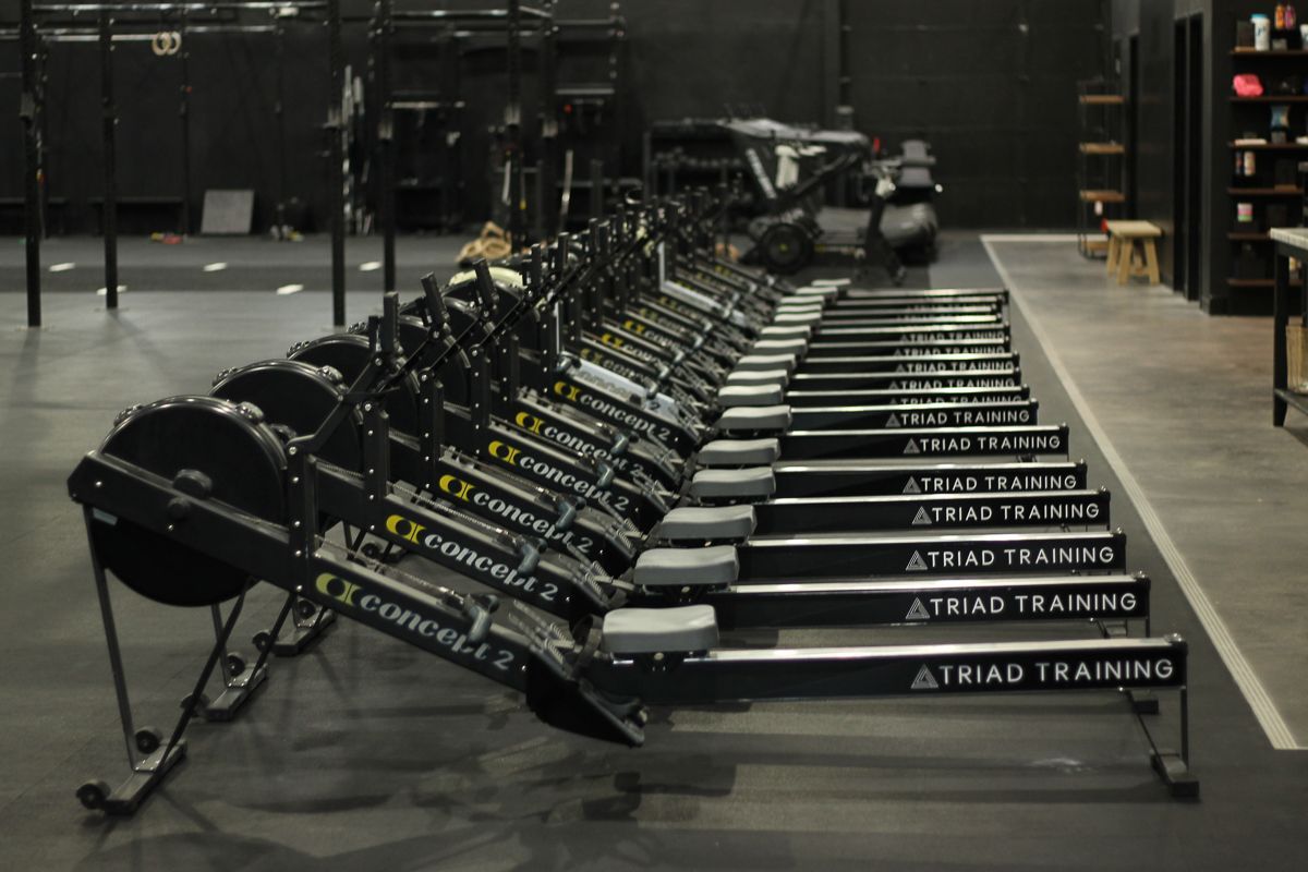 Rowing machines lined up in a gym, ready for exercise. Black and gray equipment on a dark floor.