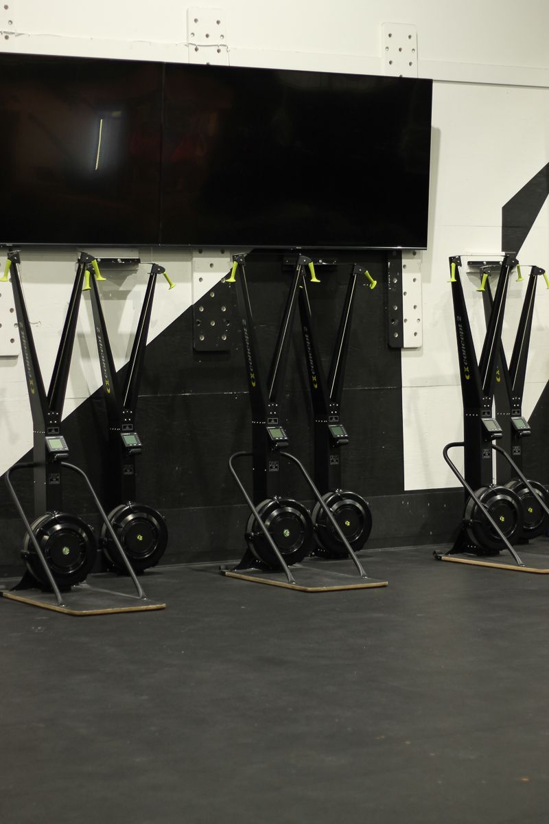 Rowing machines lined up against a wall in a gym, black and grey color scheme, with a large TV screen.