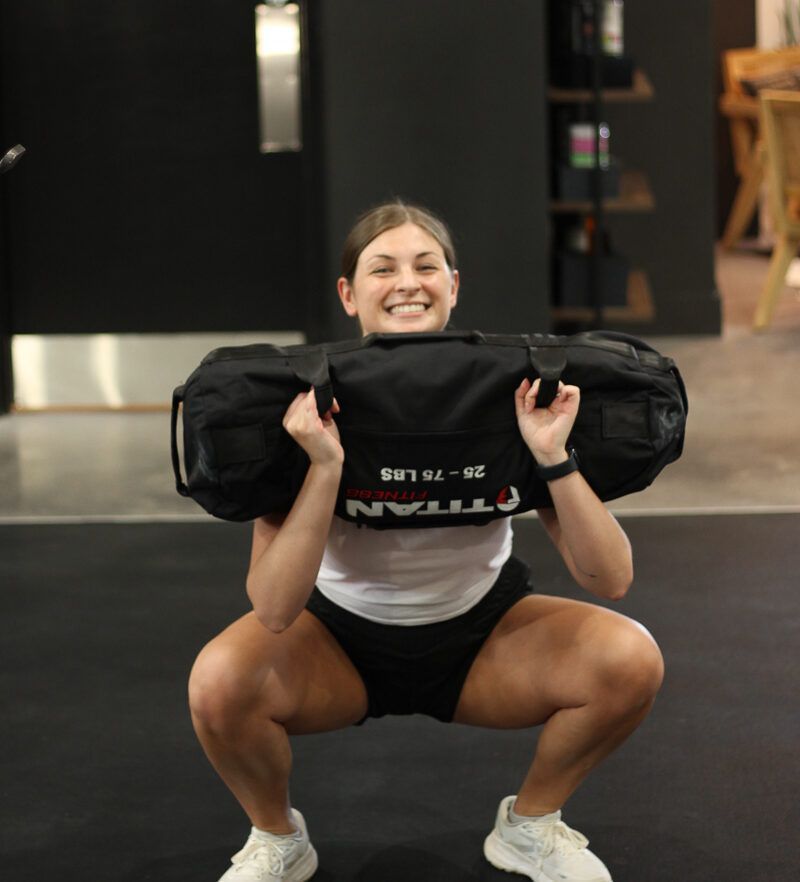 Woman in white top and black shorts doing a squat, holding a black weight bag. Gym setting.