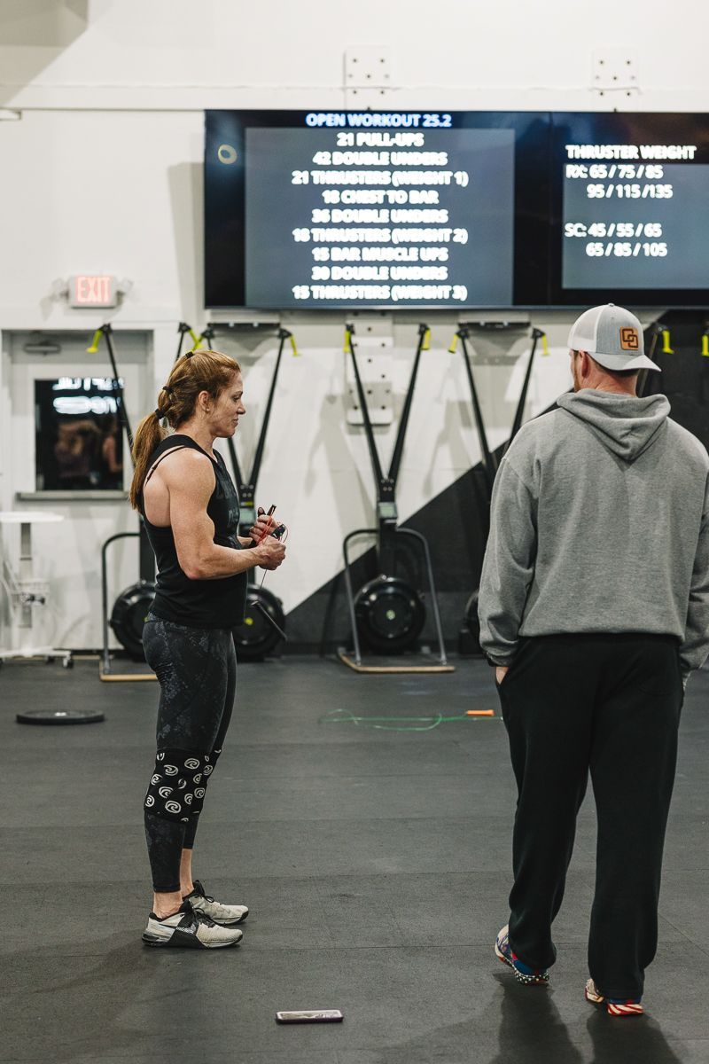 Woman in workout clothes with a coach in a gym, with workout instructions on a screen.