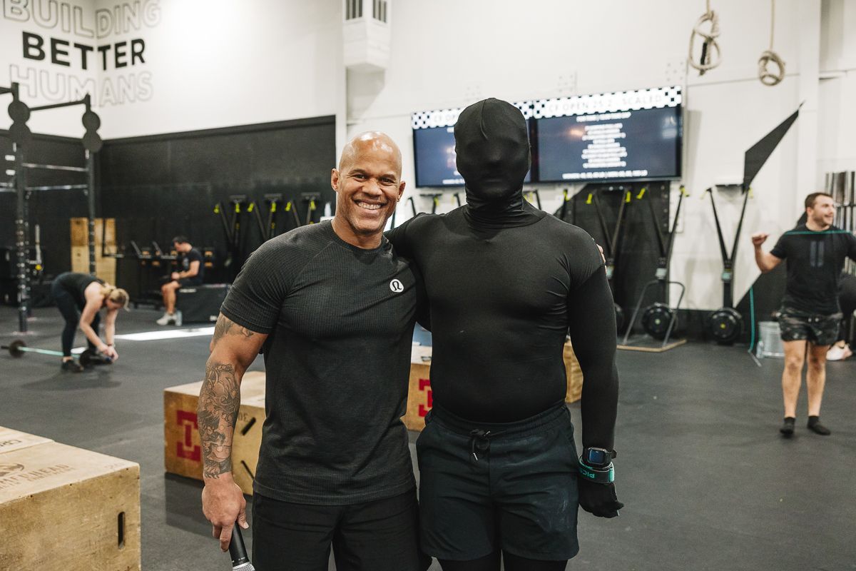 Two men in a gym pose for a photo. One smiles in a gray shirt, the other wears a full black body suit.