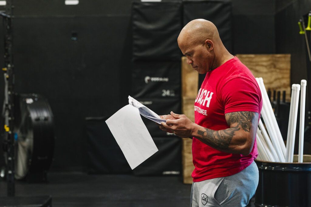 Man in red shirt looking at papers in a gym. He has tattoos and is standing near exercise equipment.
