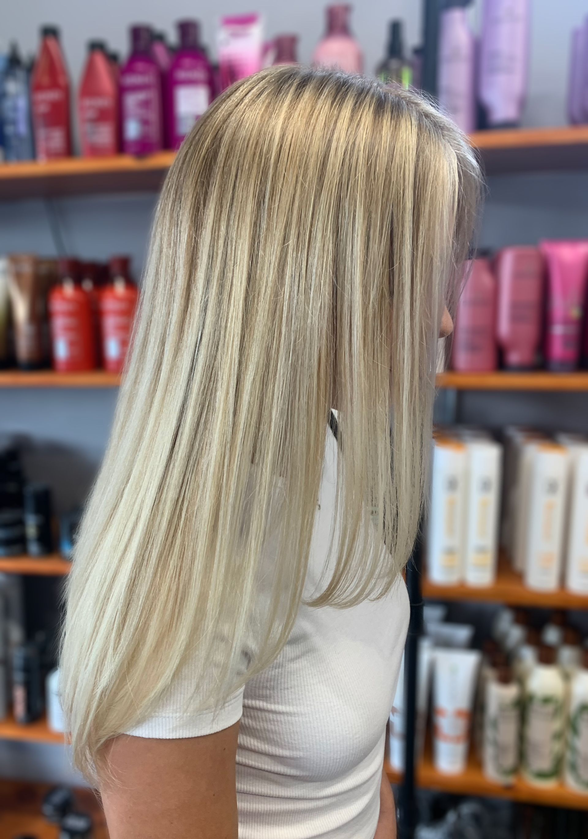 A person with long, crimped blonde hair in a salon with shelves of hair products in the background.