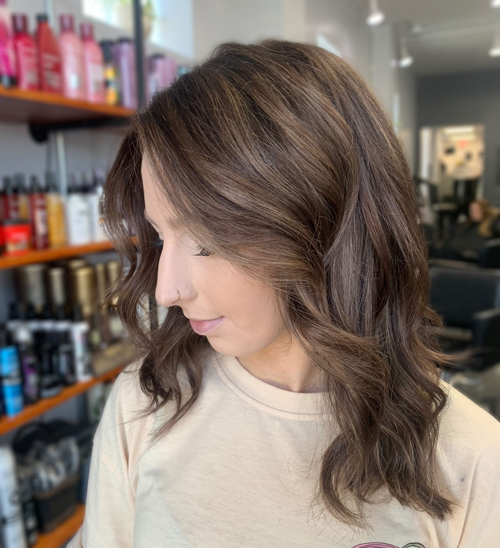 A side profile of a person with wavy, brown textured hair in a salon setting with shelves of hair products in the background.