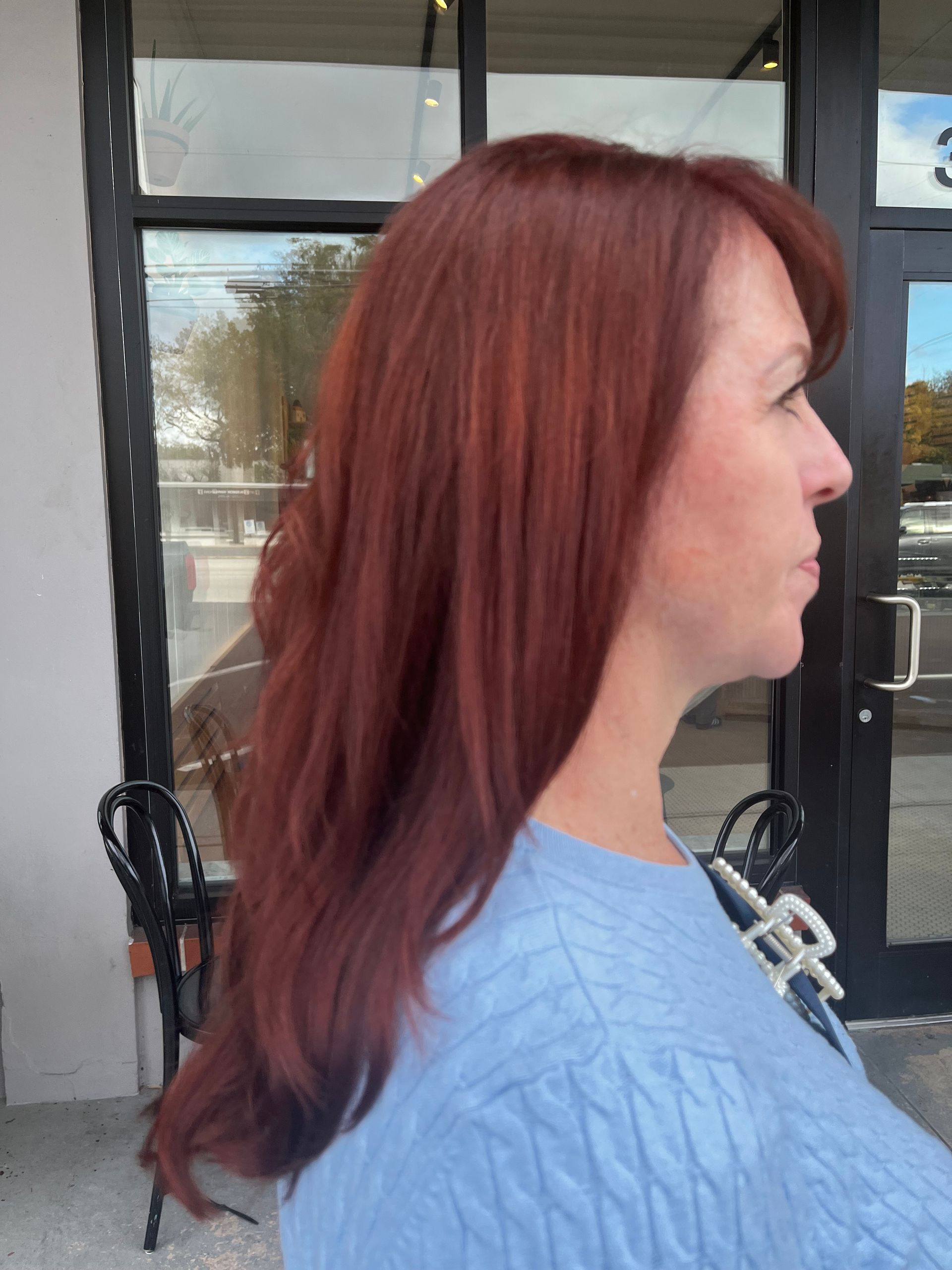 Side profile of a person with shoulder-length, deep red hair, wearing a light blue sweater, standing outside a building.