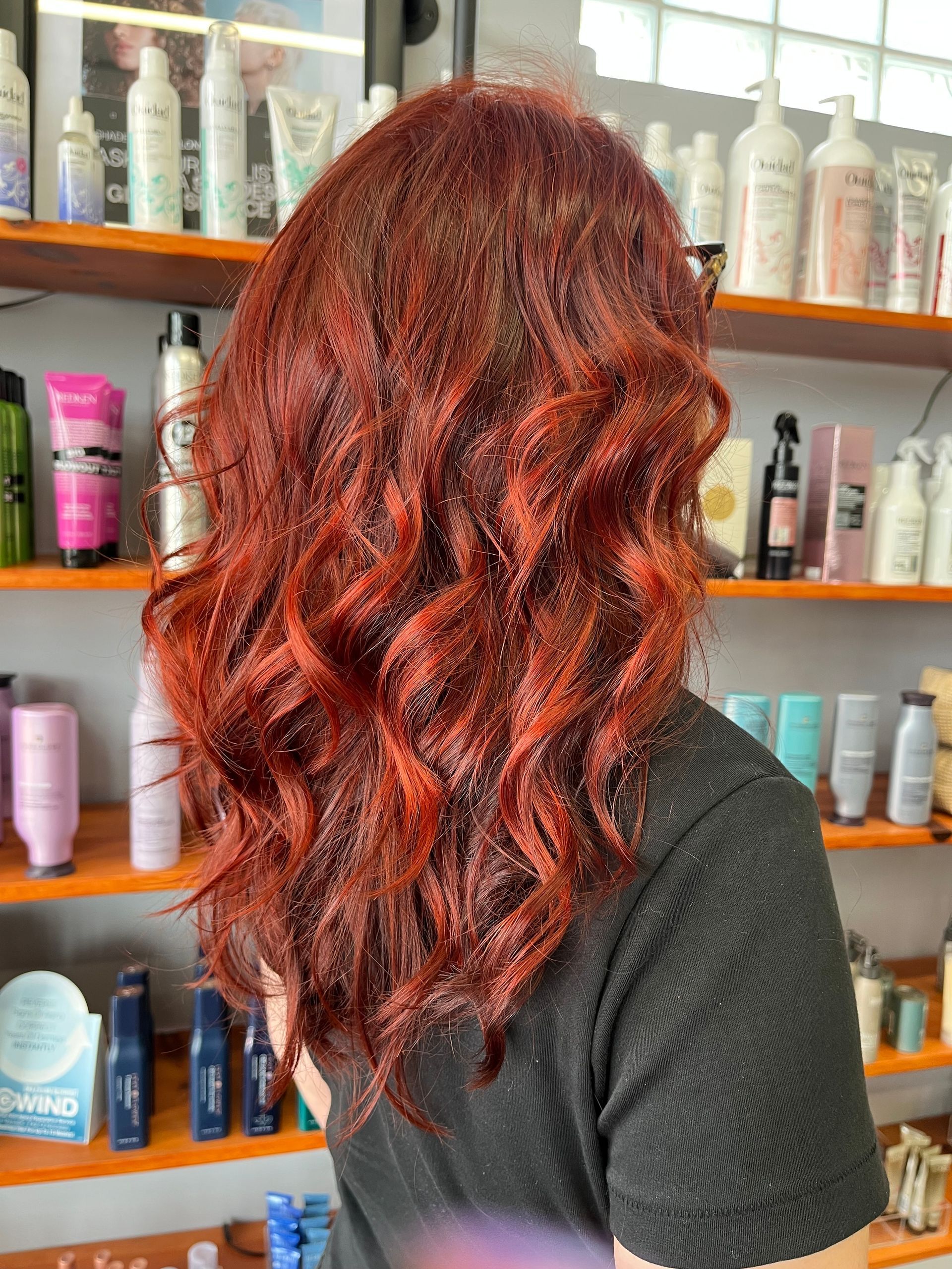 A person with shoulder-length, vibrant copper-red wavy hair, viewed from behind, standing in a hair salon.