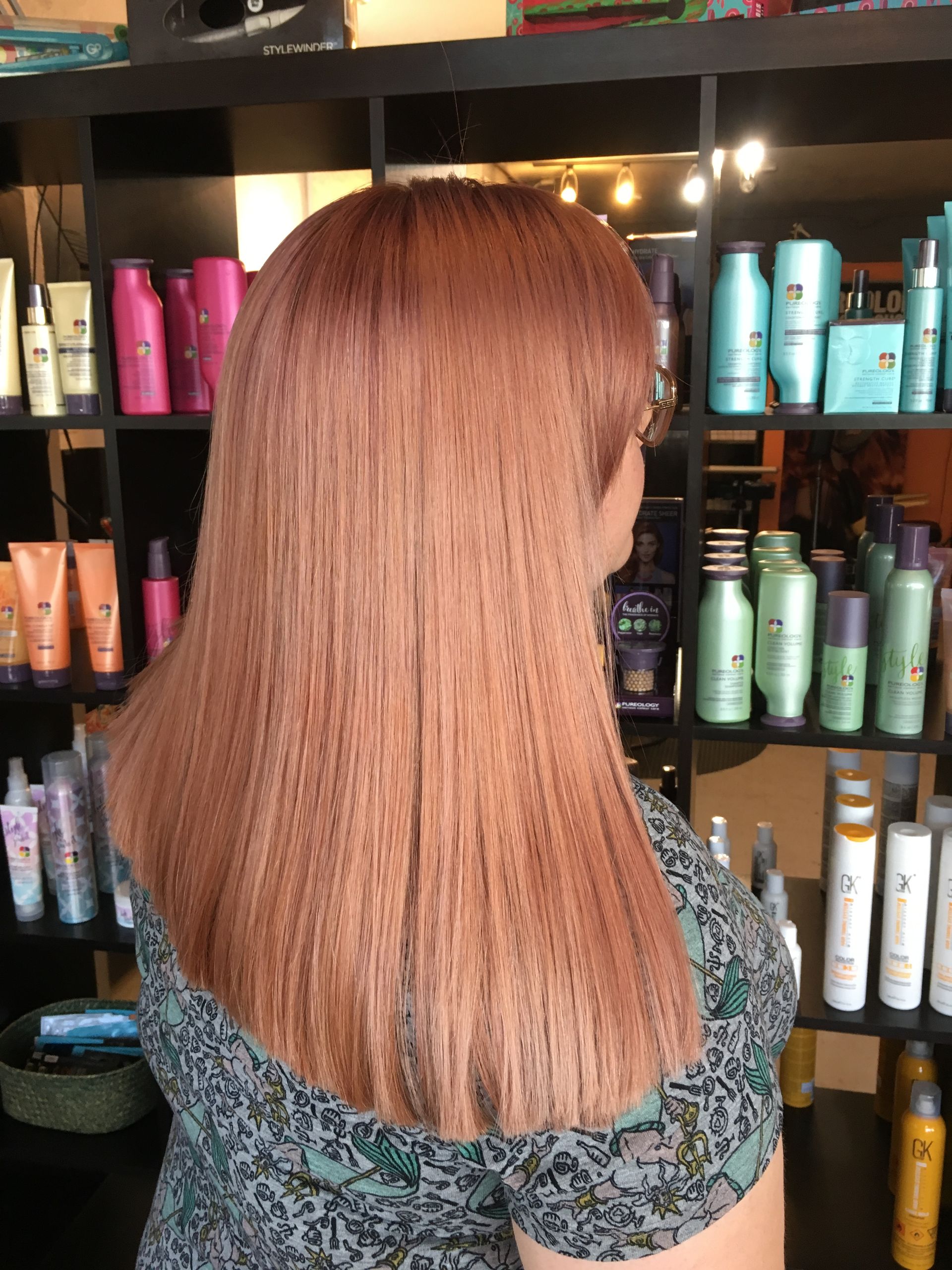 A person with shoulder-length, crimped, rose-gold hair stands in front of a shelf filled with hair product bottles.
