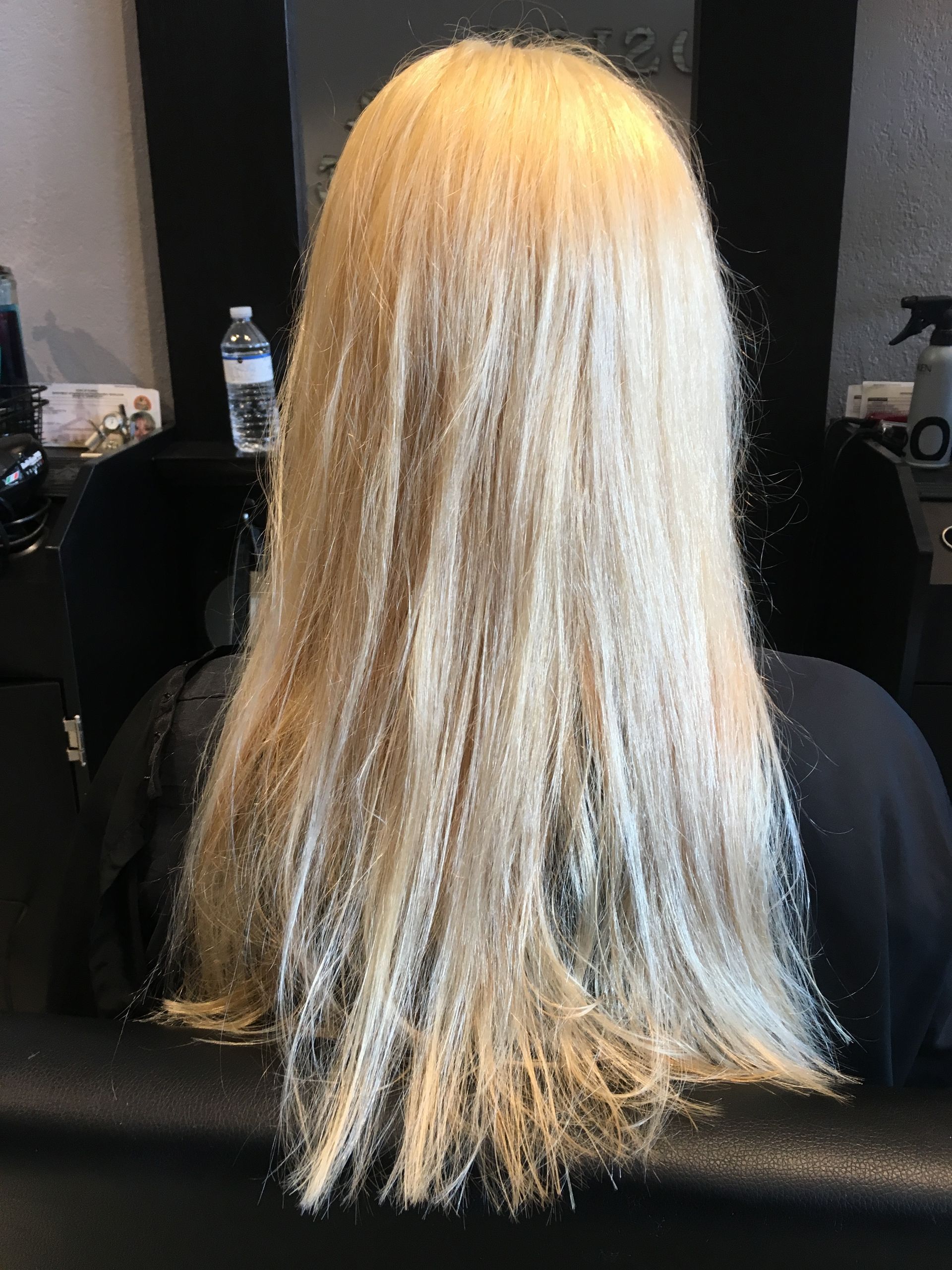A person with long, shoulder-length blonde hair that appears dry, frizzy, and damaged, seen from behind in a salon setting.