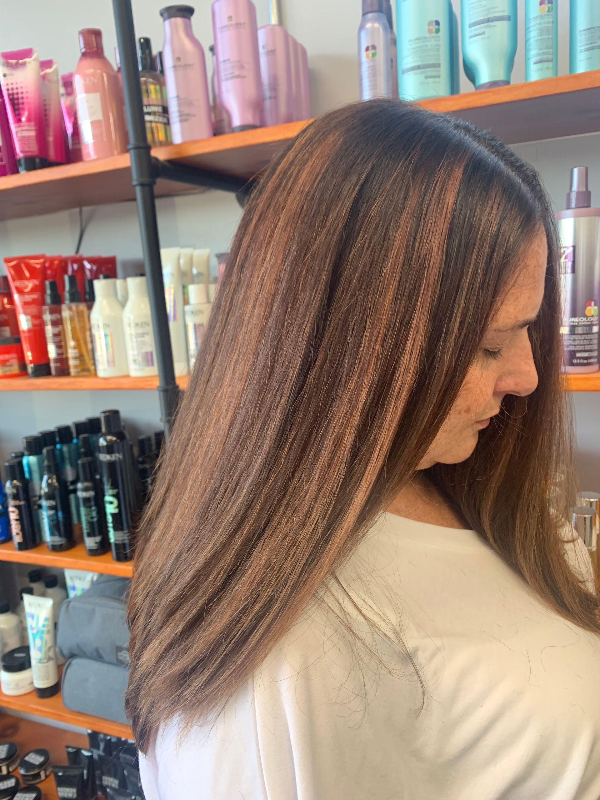 A person with long, straight, brown hair featuring copper-toned highlights, positioned in front of shelves of hair products.