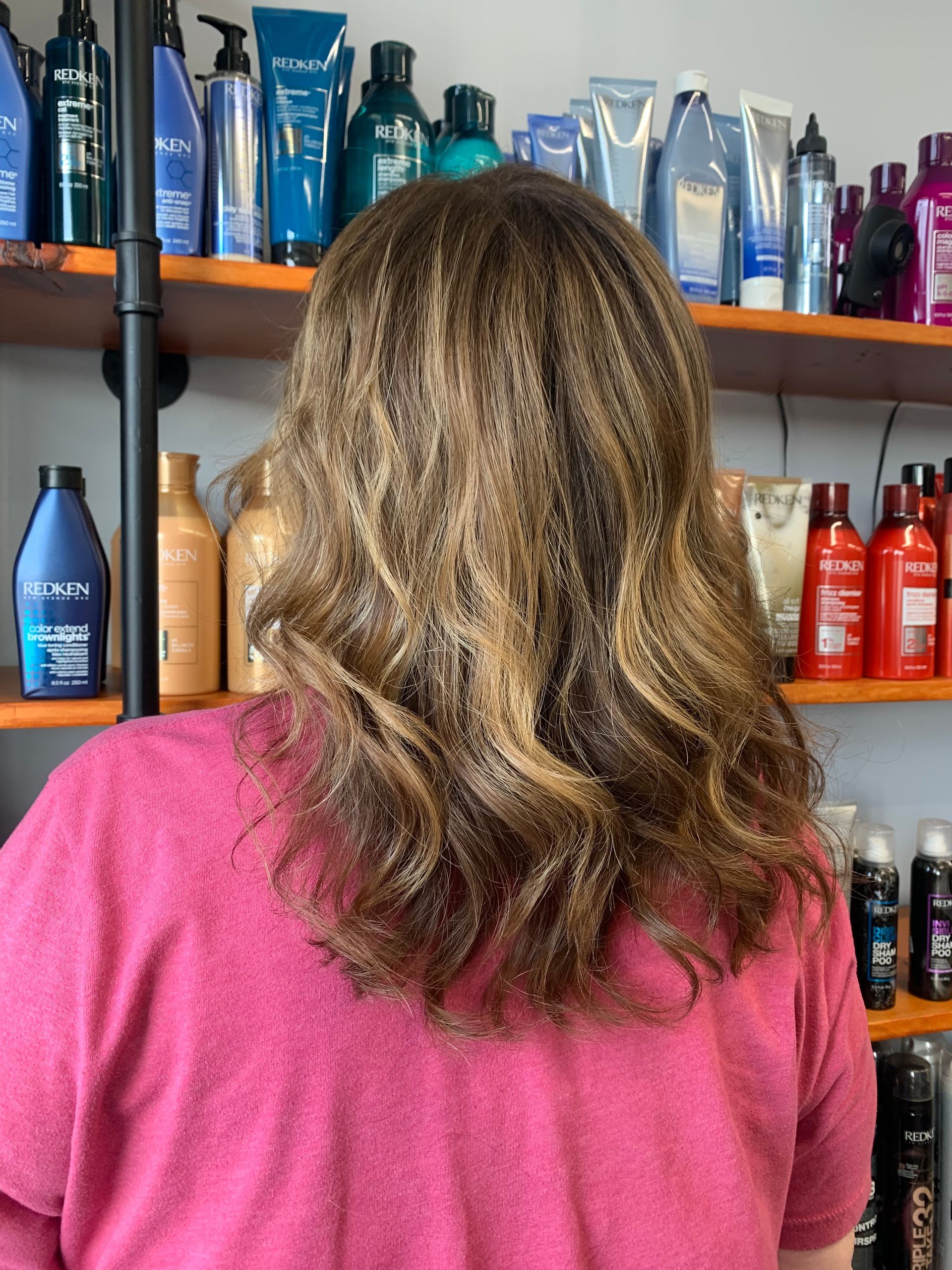 A person with wavy, highlighted brown hair stands in front of store shelves filled with various hair care products.