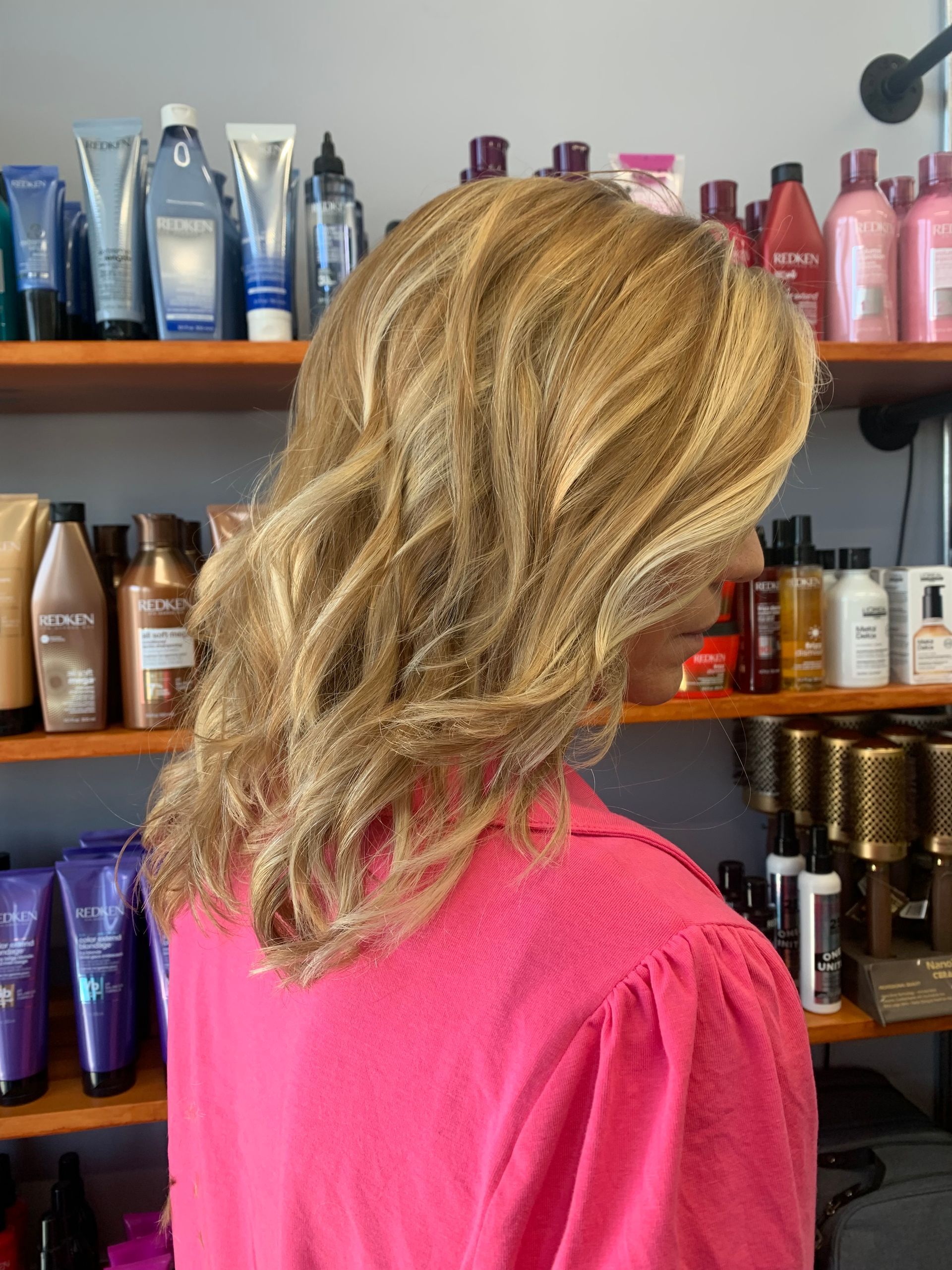 A person with wavy, highlighted blonde hair wears a bright pink top in a salon with shelves of hair care products behind.