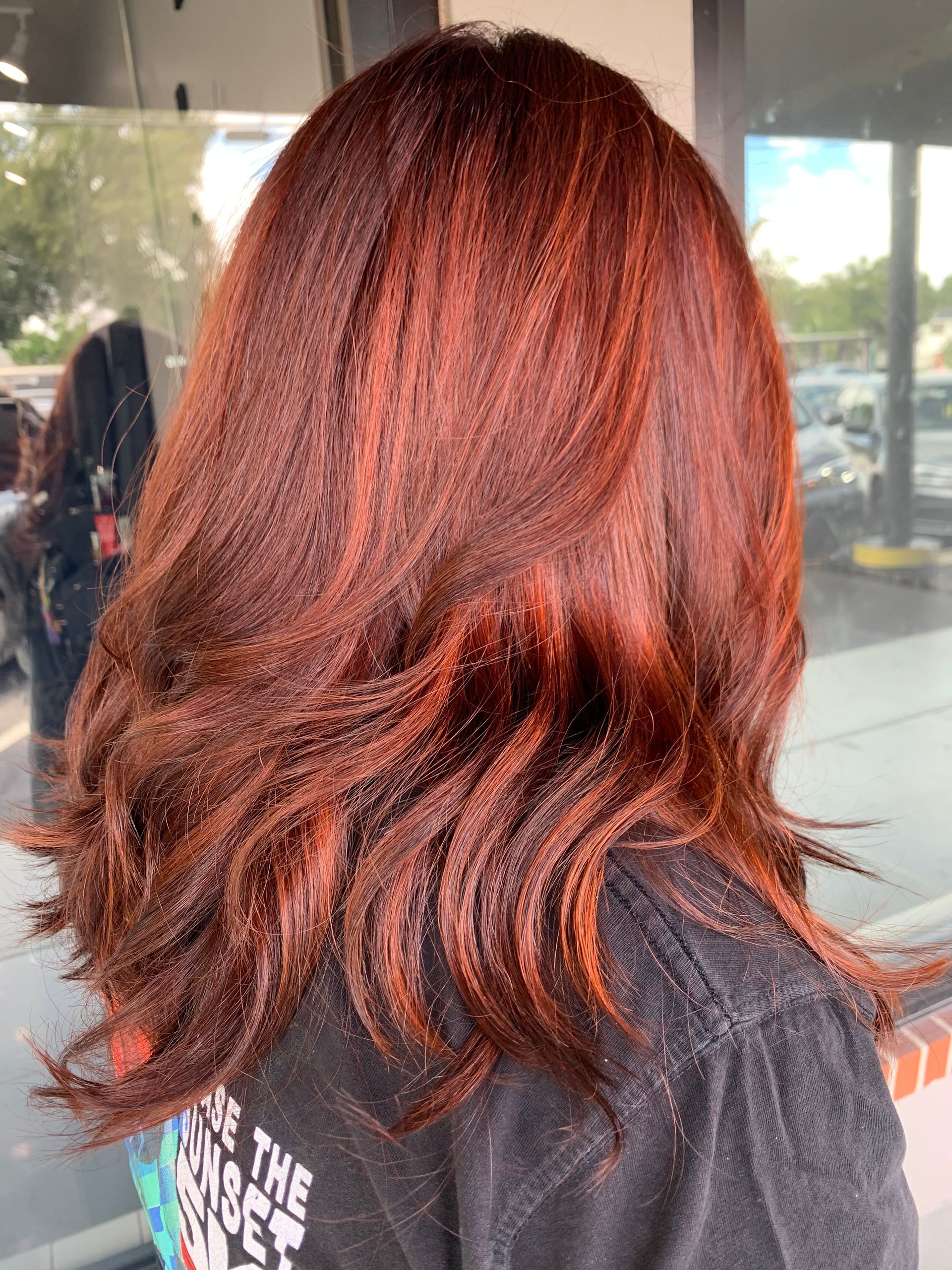 A back view of shoulder-length, textured hair dyed a deep auburn red, styled with loose, flowing waves.