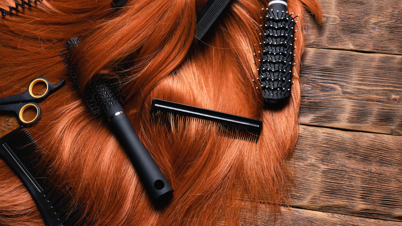A collection of hair styling tools, including scissors, combs, and a round brush, resting on vibrant copper-colored hair.