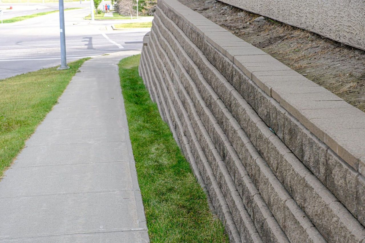 A sidewalk runs alongside a tiered concrete retaining wall with a grassy strip in between, set in an outdoor urban area.
