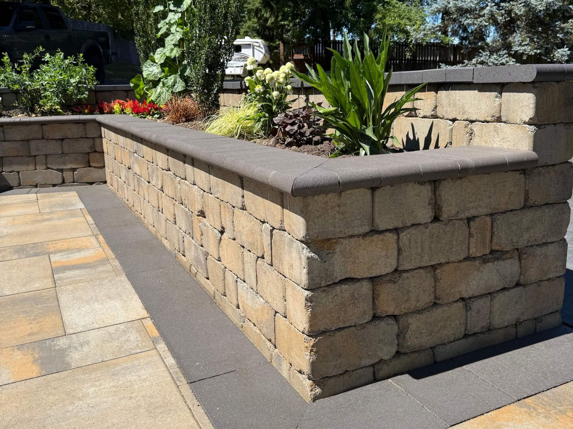 A beige stone retaining wall with a dark grey cap, bordering a patio with plants growing in the raised garden bed.