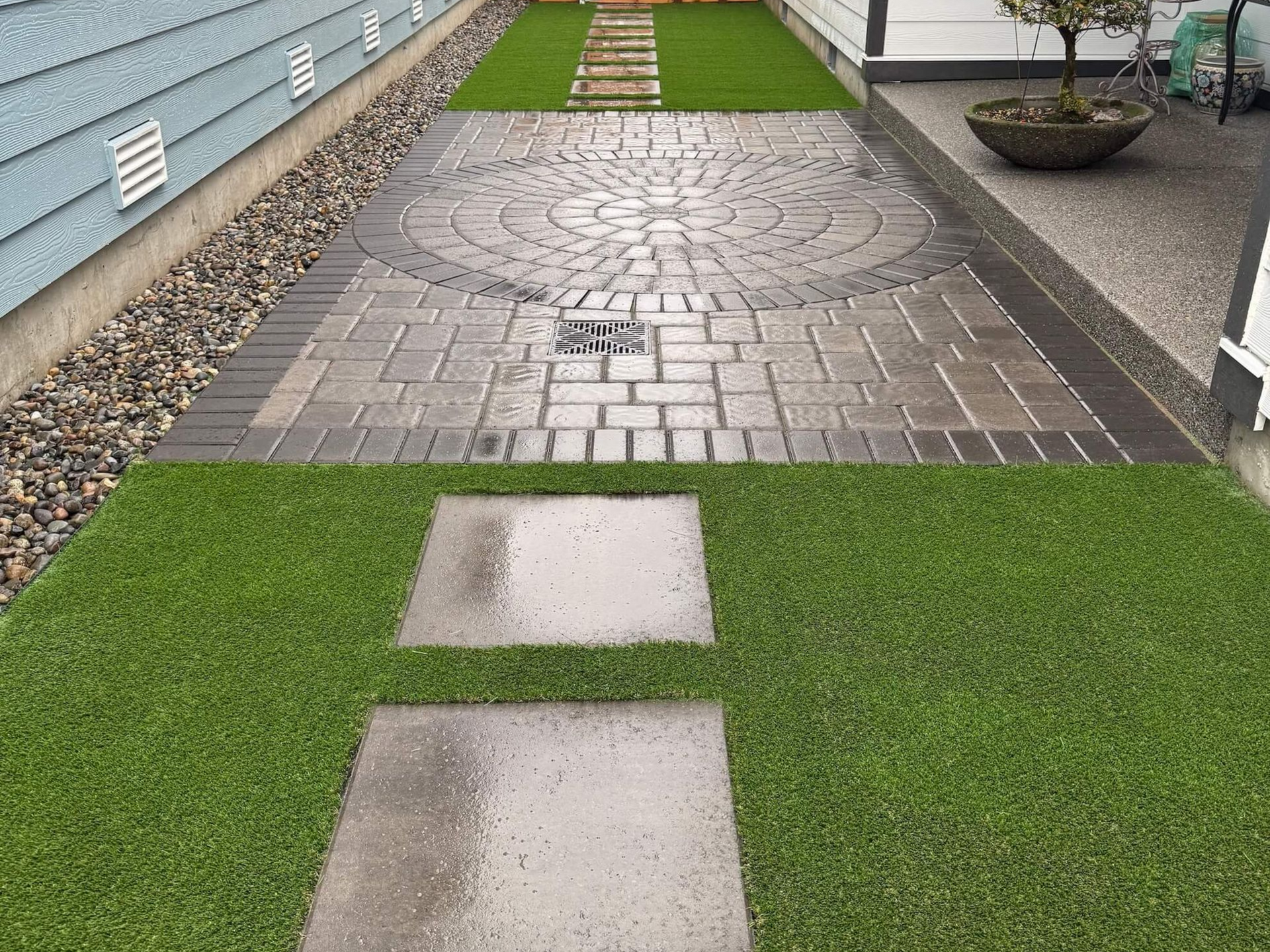 A paved backyard patio with a stone circular design, surrounded by artificial grass and a rock border between two houses.