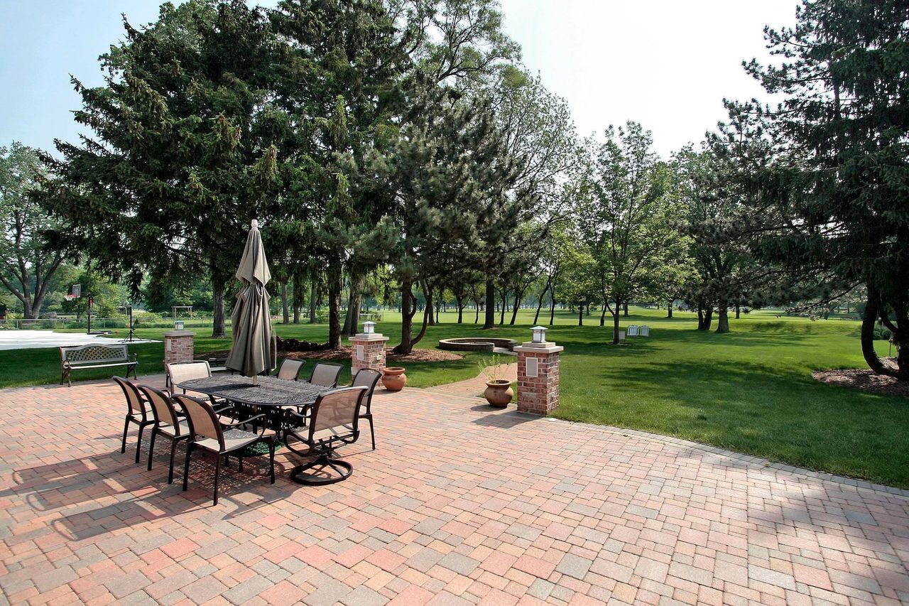 Patio with a metal dining set and umbrella on a red brick floor, overlooking a lush green lawn and mature trees.