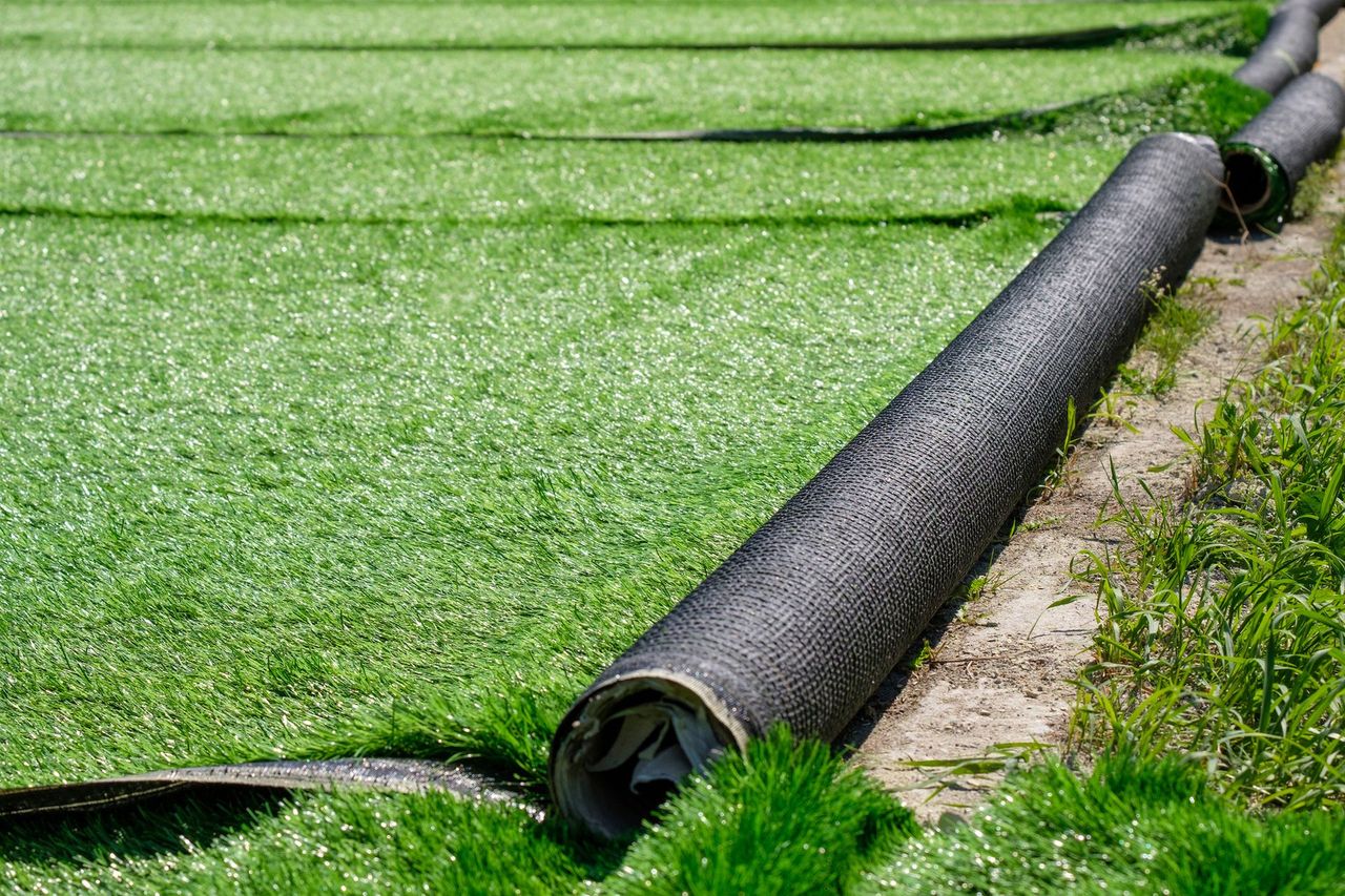 A roll of green artificial grass partially unrolled onto a dirt surface with other strips of turf laid out in the background.