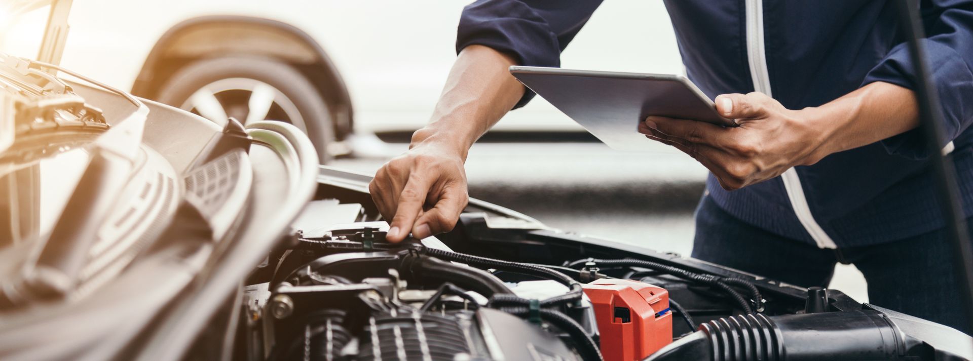 A mechanic is looking under the hood of a car while holding a tablet. | Halton Auto Service & Transmission