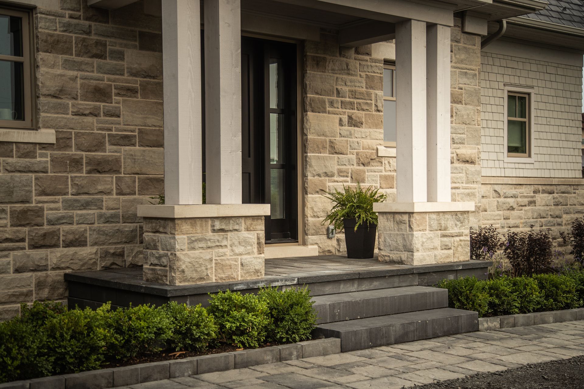 Stone facade house with dark front door, white pillars, and stone steps. Green plants in planters and a garden.