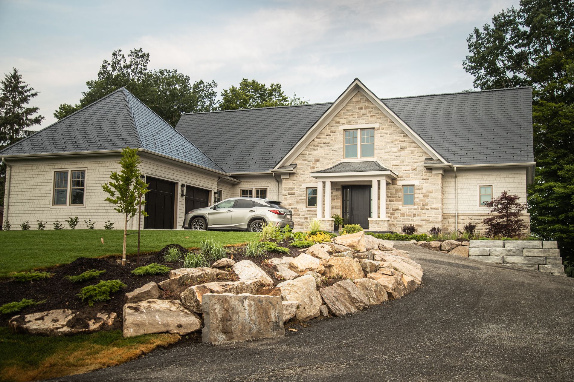 Stone-clad house with a dark gray roof, driveway, and rock landscaping. A car is parked in the garage.