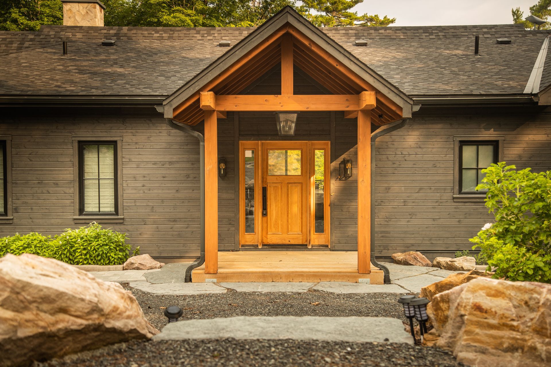 Wooden-framed entrance to a house with gray siding, light wooden door under a gabled roof. Stone steps lead up.