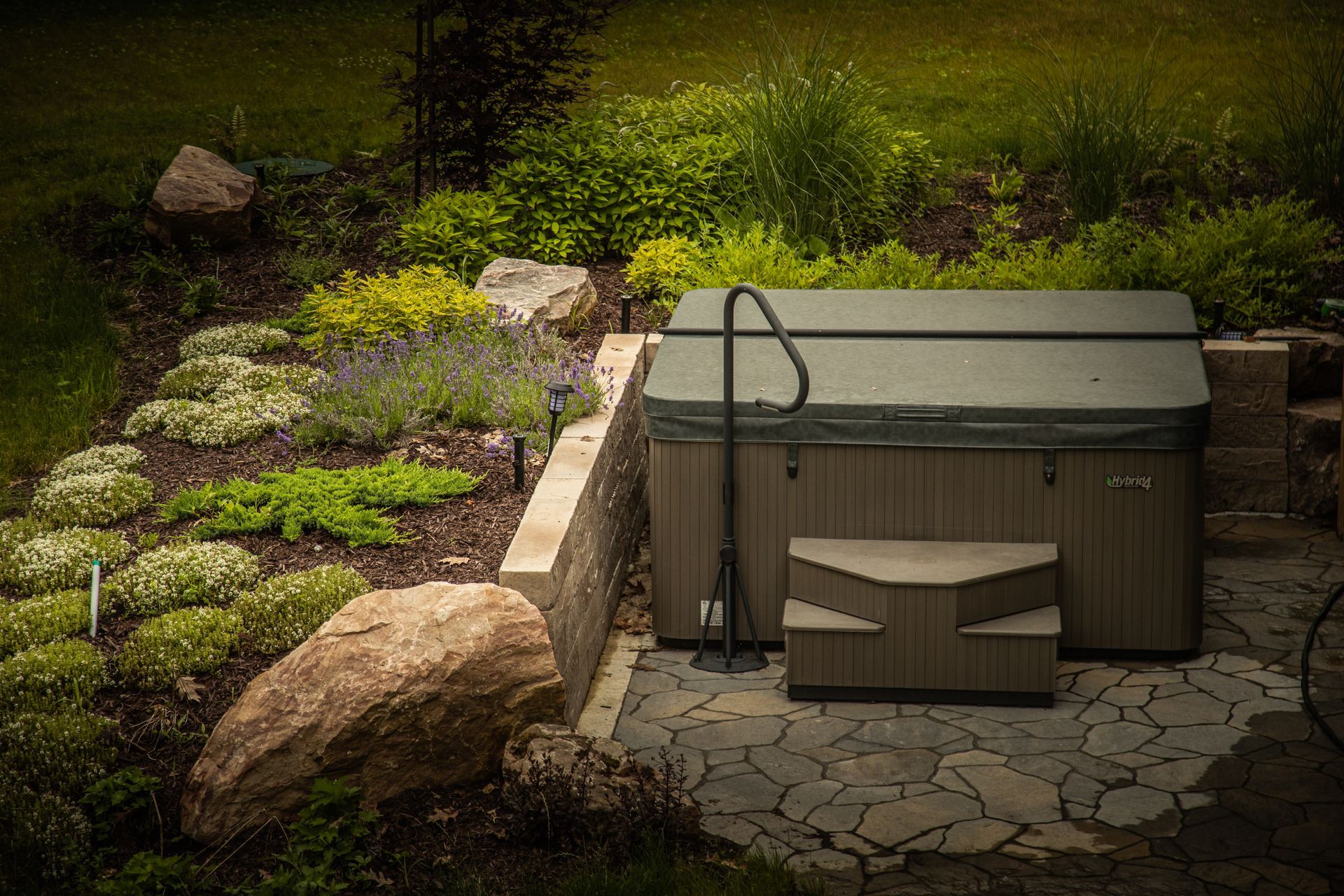 Hot tub with cover on stone patio, surrounded by landscaping, in outdoor setting.
