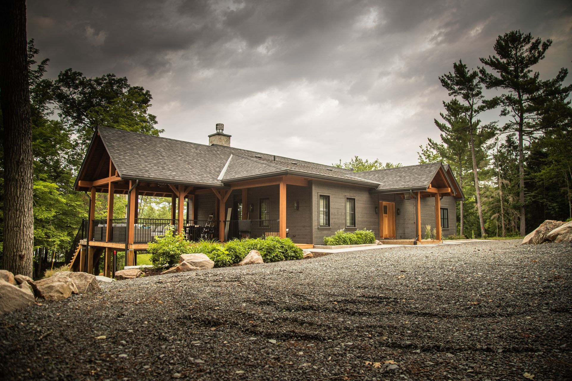 A dark-grey modern house with a wooden porch and a gravel driveway under a cloudy sky.
