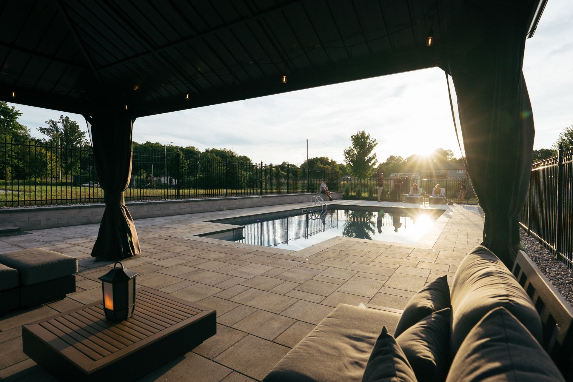 Outdoor patio with pool. Gazebo with draped curtains. Cushioned seating, lantern on table. Sunlit pool and sky.