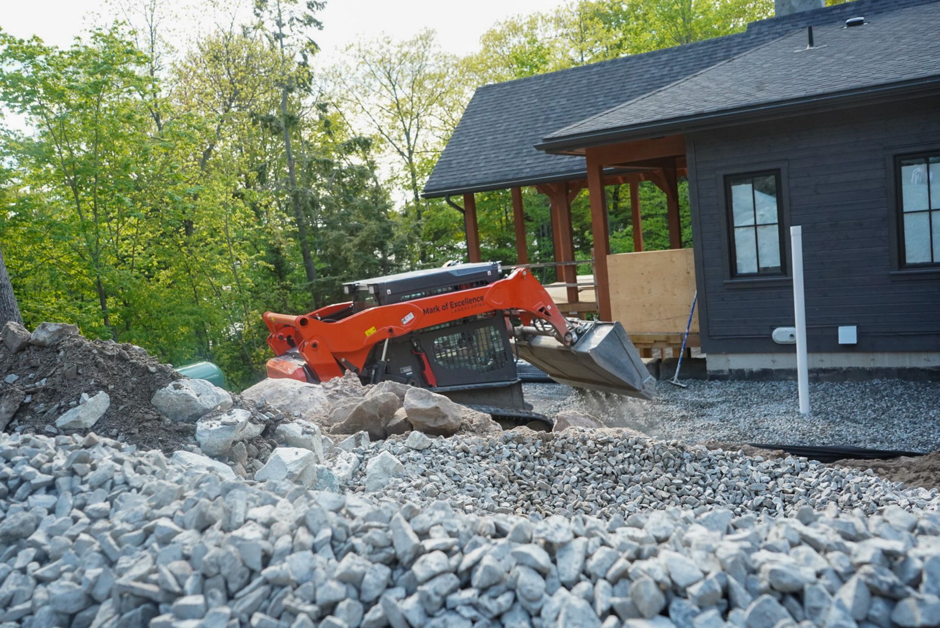Orange skid steer moving gravel near a house with a wooden porch and dark siding.
