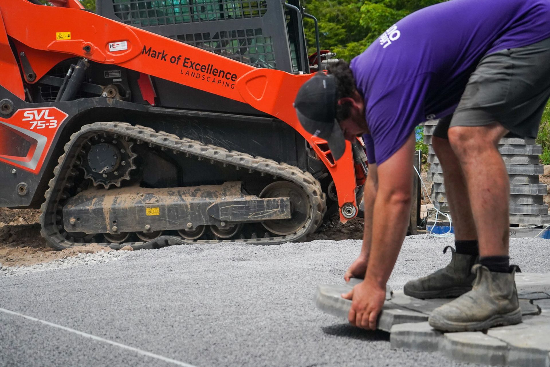 Man in purple shirt placing pavers next to an orange skid steer on a gravel surface.