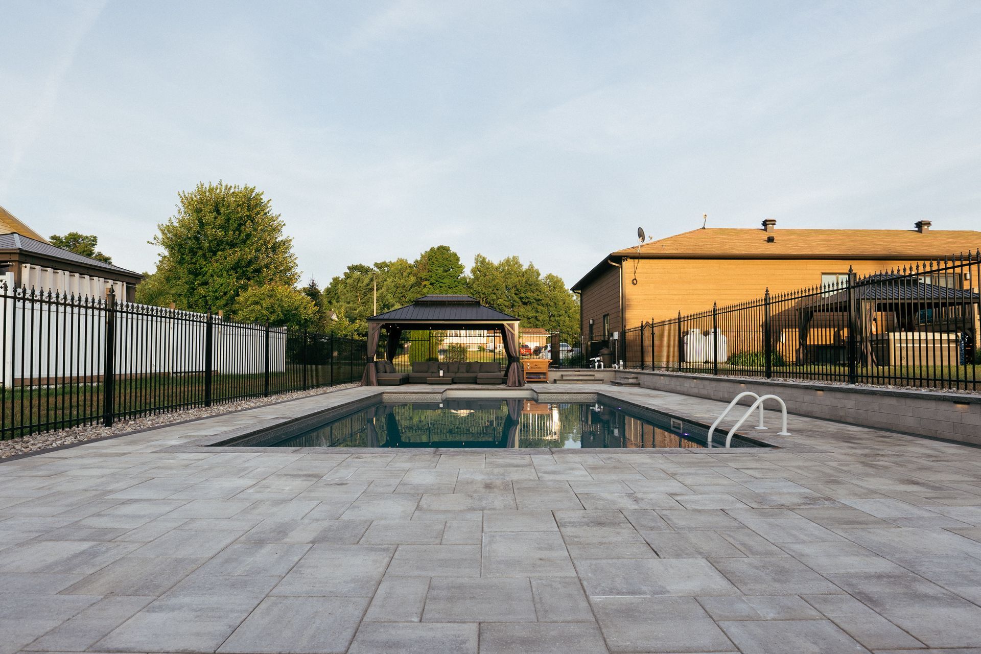 Rectangular pool surrounded by gray paving, fenced yard, gazebo, and yellow brick building.