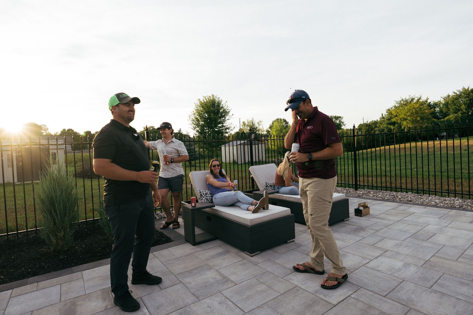 Four people relaxing on a patio at sunset; two men standing, two on lounge chairs, with drinks.