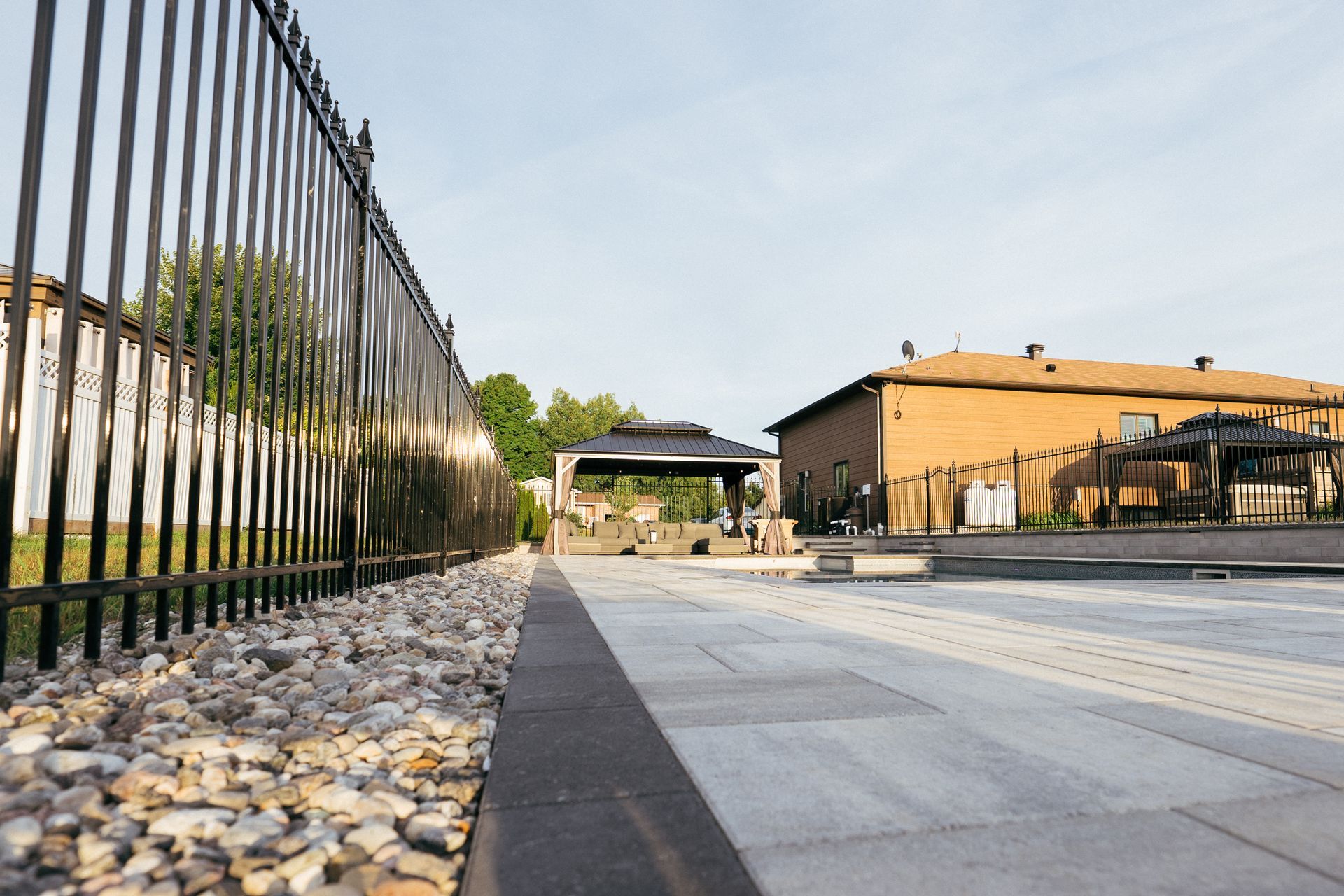 Black metal fence alongside a paved walkway with a gravel border, leading toward a gazebo and brick house under a blue sky.