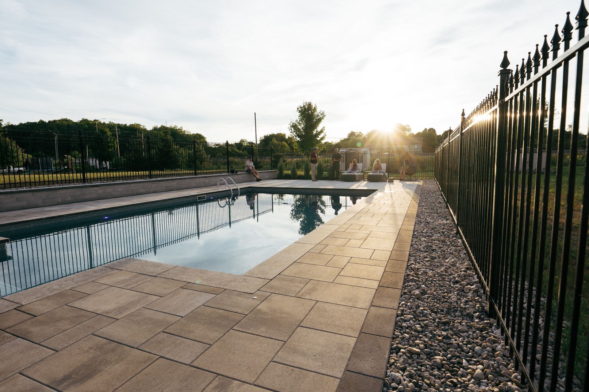 Poolside view with a rectangular pool, pavers, black fence, and trees at sunset.