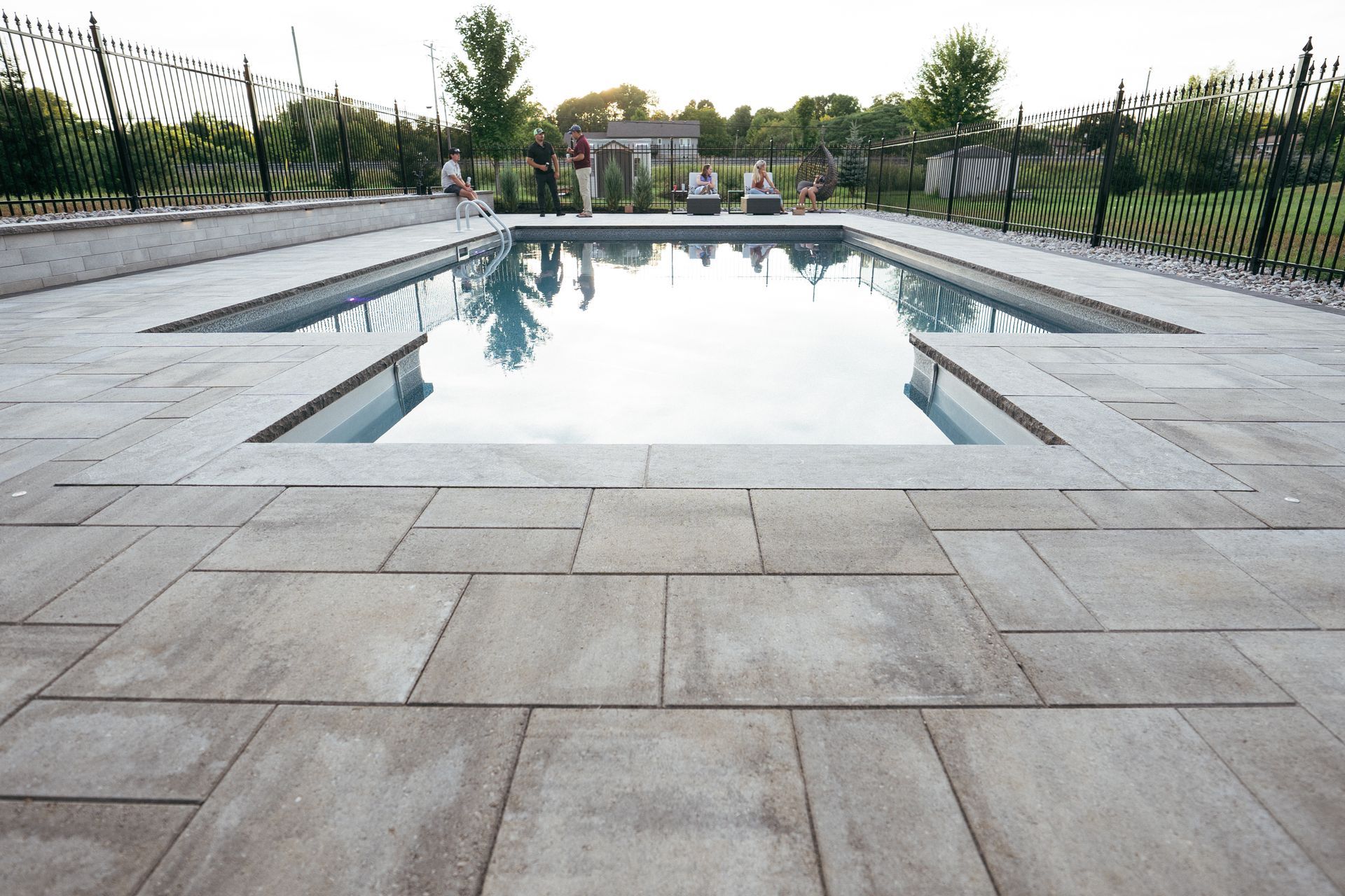 Rectangular pool with gray pavers, black fence, and people in the background.