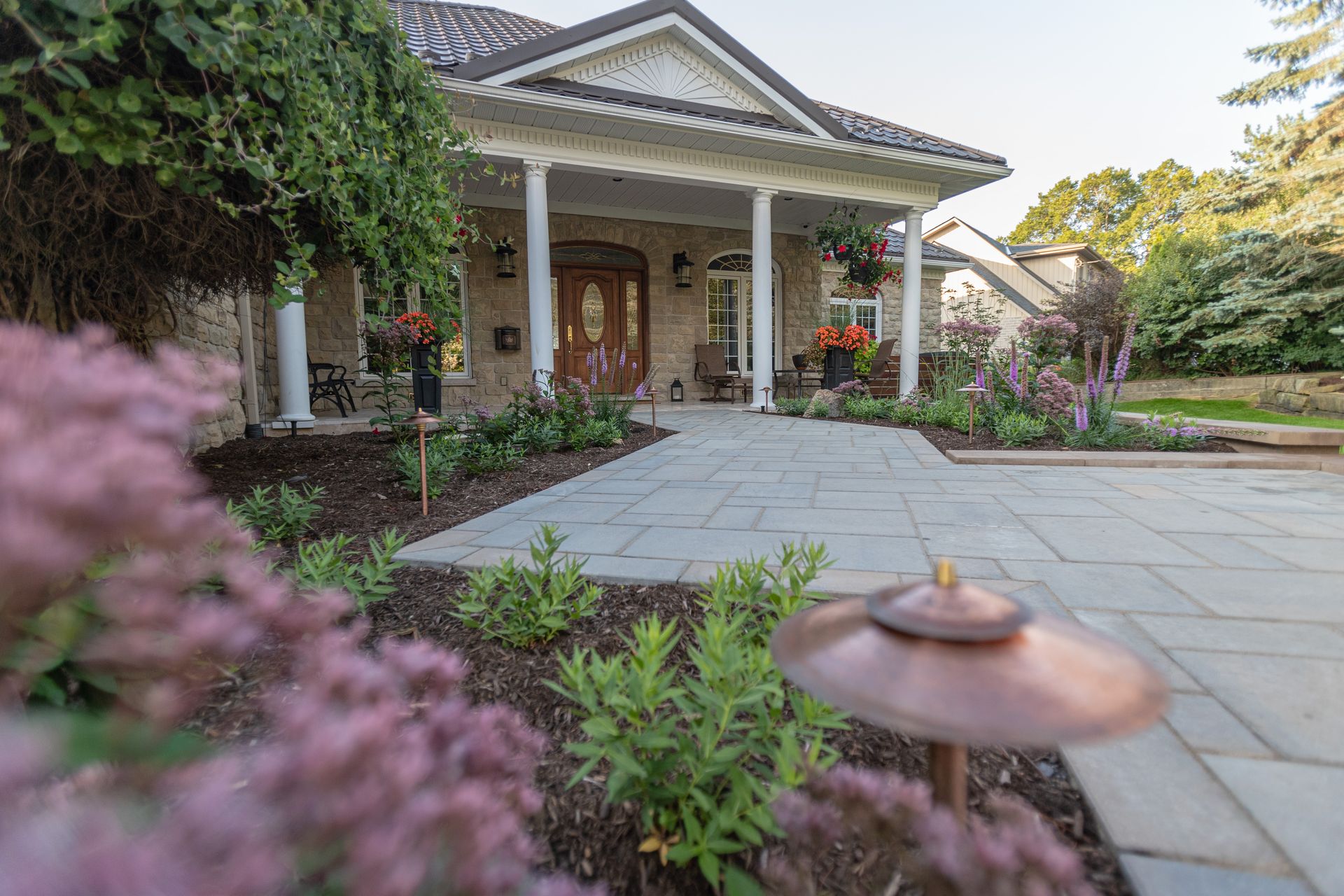 Stone walkway leading to a house with a covered porch. Landscaping includes flowers and a copper-colored path light.