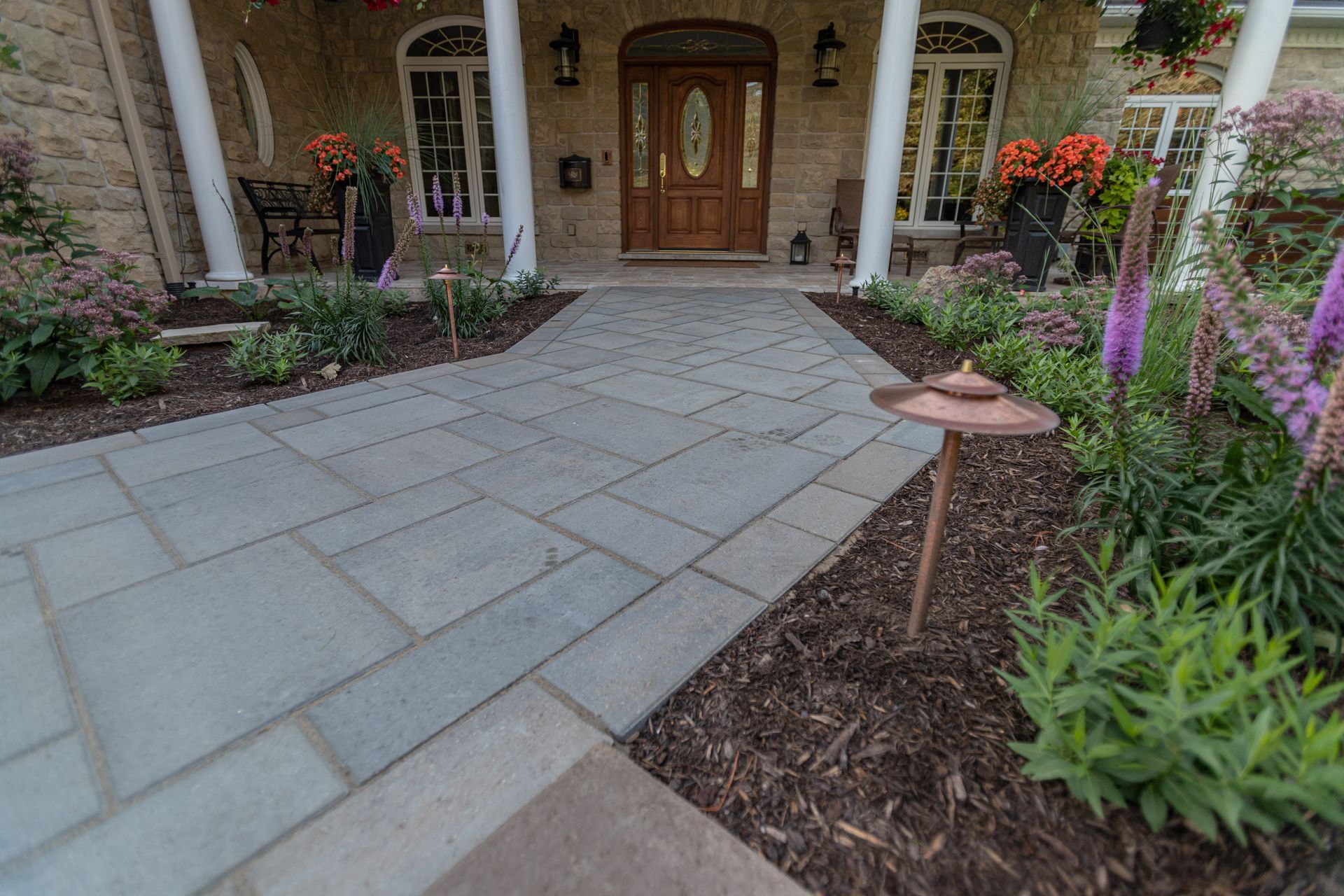 Stone walkway leading to a house's wooden door with landscaping. Bronze lights border the path.